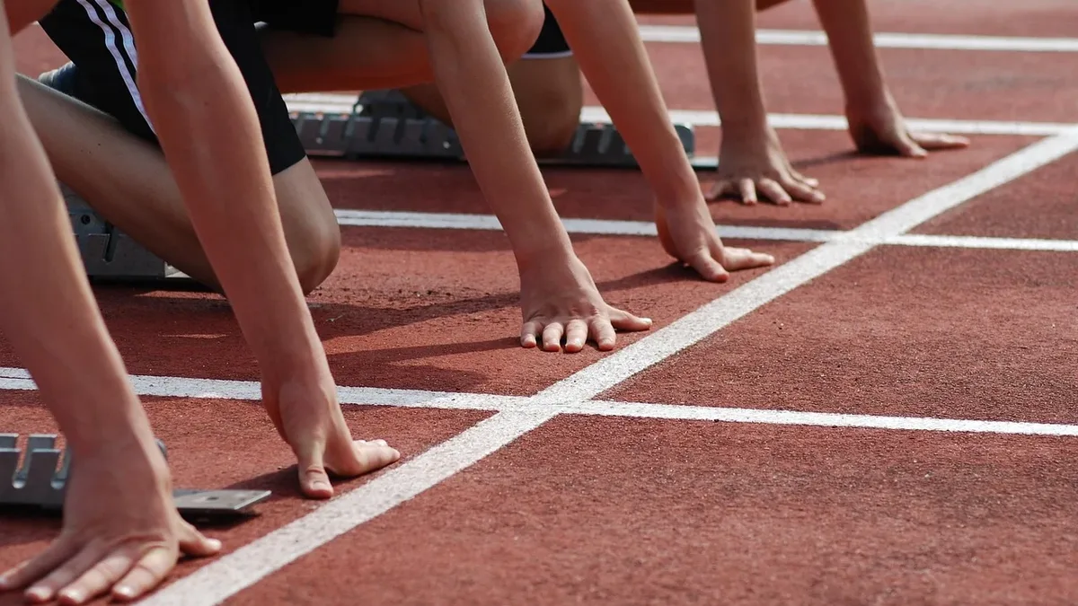The legs and feet of several people lined up at the starting line of a track, with their toes on the starting blocks.