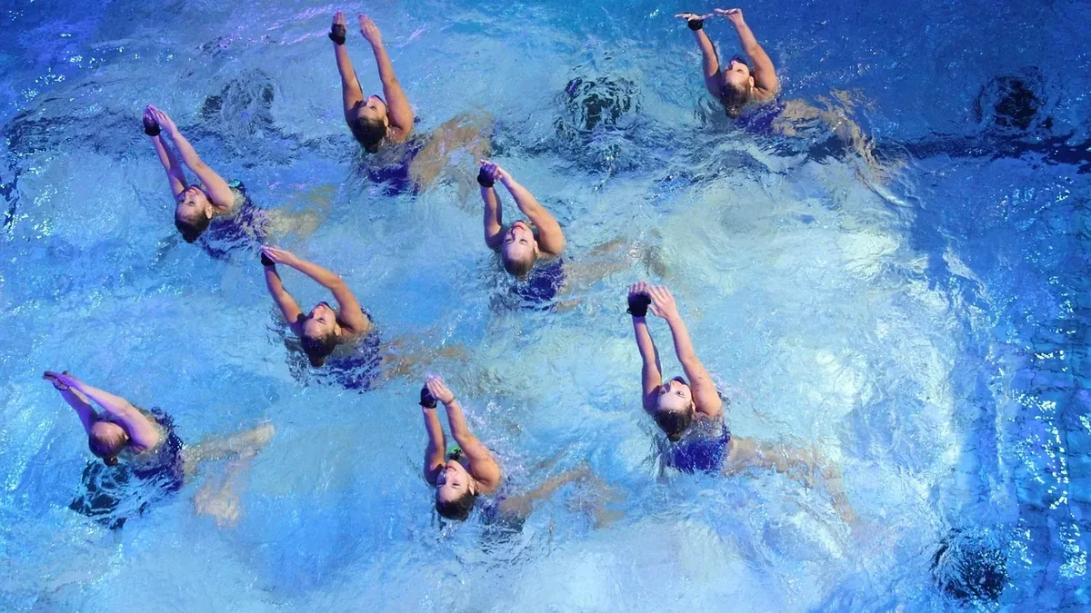 Eight synchronized swimmers are performing in a pool, with their bodies positioned in a circular formation and their arms extended above the