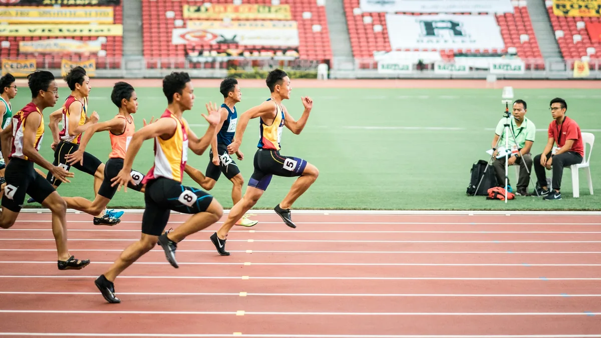 A group of young men in athletic uniforms are running on a track, with a man sitting on a chair in the background.